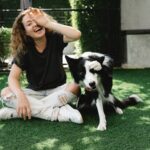 Cheerful female covering eye while sitting with crossed legs against purebred dog and looking at camera on meadow in sunlight