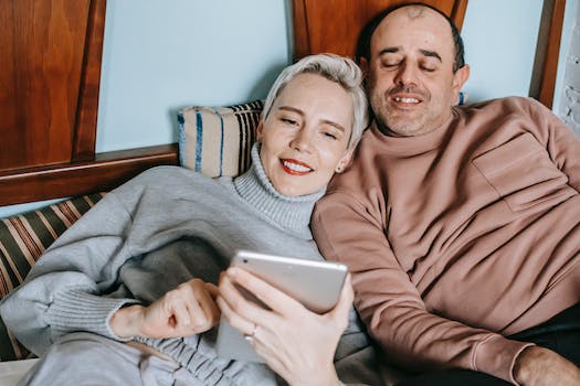 Content diverse middle aged married couple in warm casual clothes lying together on bed and smiling while watching movie on tablet