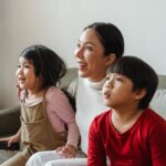 Cute little Asian kids watching exciting movie with mom while resting on sofa at home