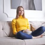 Positive young female in casual clothes sitting on cozy sofa with bowl of snacks and watching interesting film while spending time at home
