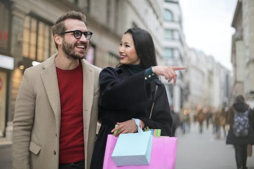 From below happy stylish Asian woman in warm clothes smiling at cheerful unshaven man in glasses and pointing finger away while walking along street together in city after shopping