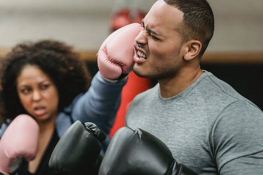 Side view of young muscular ethnic male trainer in sportswear and boxing gloves receiving heavy punch on face from serious young African American female during workout