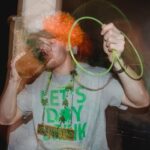 Young male in clown party wig colored in Ireland national colors drinking mug of beer with neon glowstick necklace celebrating St Patrick Day