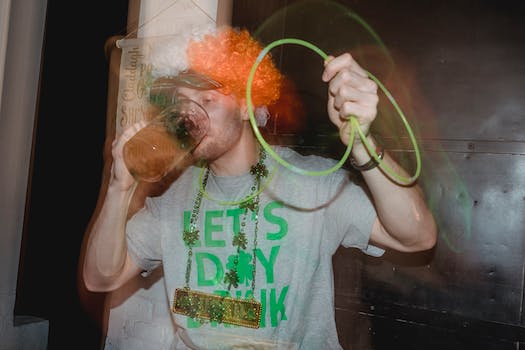 Young male in clown party wig colored in Ireland national colors drinking mug of beer with neon glowstick necklace celebrating St Patrick Day