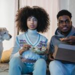 Concentrated young African American couple with curly hairs in casual outfits eating takeaway salad and pizza while watching TV sitting on sofa near cute purebred dog