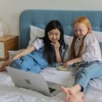 Full body of happy barefooted diverse children with long hairs lying on comfortable bed and eating popcorn while watching funny cartoon on laptop in cozy room
