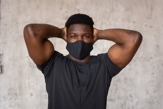 Serious African American male with short dark hair in protective mask standing with raised arms and looking at camera against concrete wall