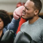 Side view of young muscular ethnic male trainer in sportswear and boxing gloves receiving heavy punch on face from serious young African American female during workout