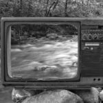 Black and white vintage old broken TV placed on stones near wild river flowing through forest
