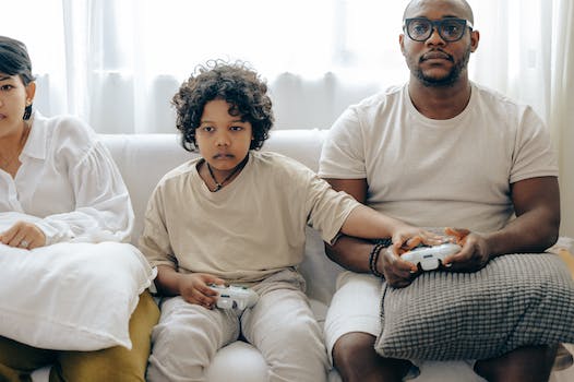Concentrated child showing usage of gamepad to father while playing on game console and resting on sofa together with family