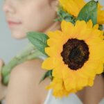 Crop anonymous woman in casual clothes holding bright sunflowers on shoulder and showing to camera against blurred background