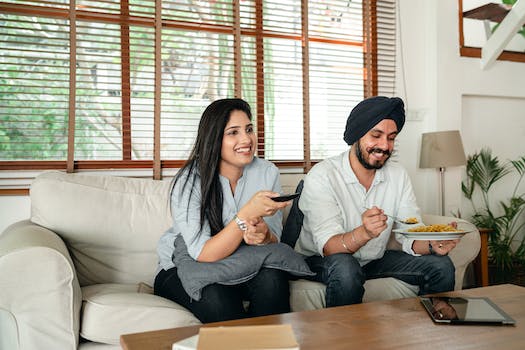 Happy ethnic male with plate of food and female with remote control sitting on couch in living room while watching movie