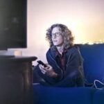 Concentrated male with long hair sitting on comfortable sofa at home and messaging on social media via cellphone while watching movie on TV with opened mouth