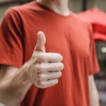 Crop anonymous young male in red t shirt showing thumb up gesture while standing on city street on sunny day