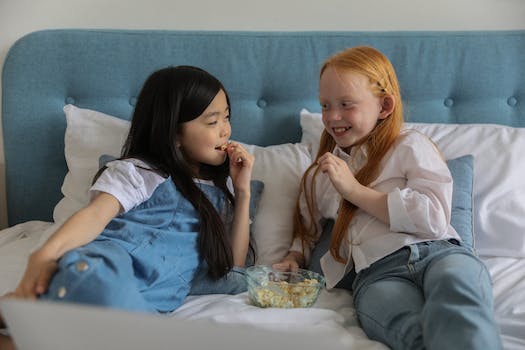 Happy little multiethnic children with dark and red hairs lying on comfortable bed and looking at each other while eating popcorn during lazy weekend