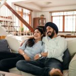 Happy young Indian man in turban and positive woman in casual clothes embracing and laughing while watching comedy movie sitting on sofa