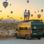 Two people standing on top of a van looking at hot air balloons